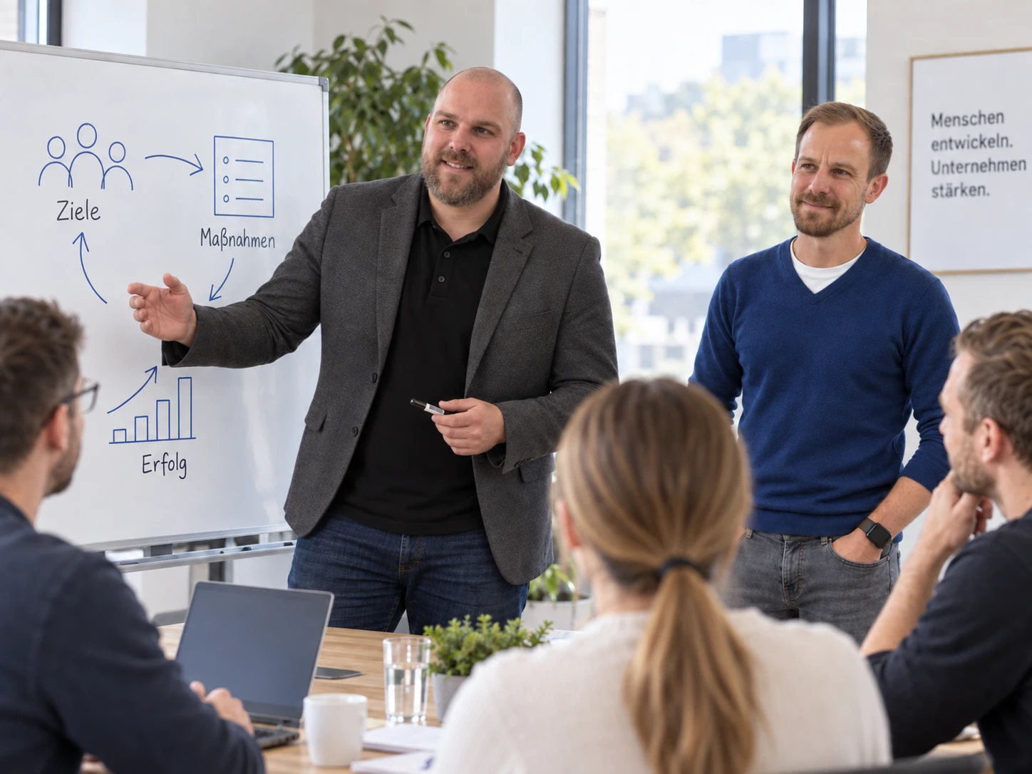 Freddy erklärt Ziele und Schritte am Whiteboard; David begleitet die Schulung. KI-generiertes Symbolbild.